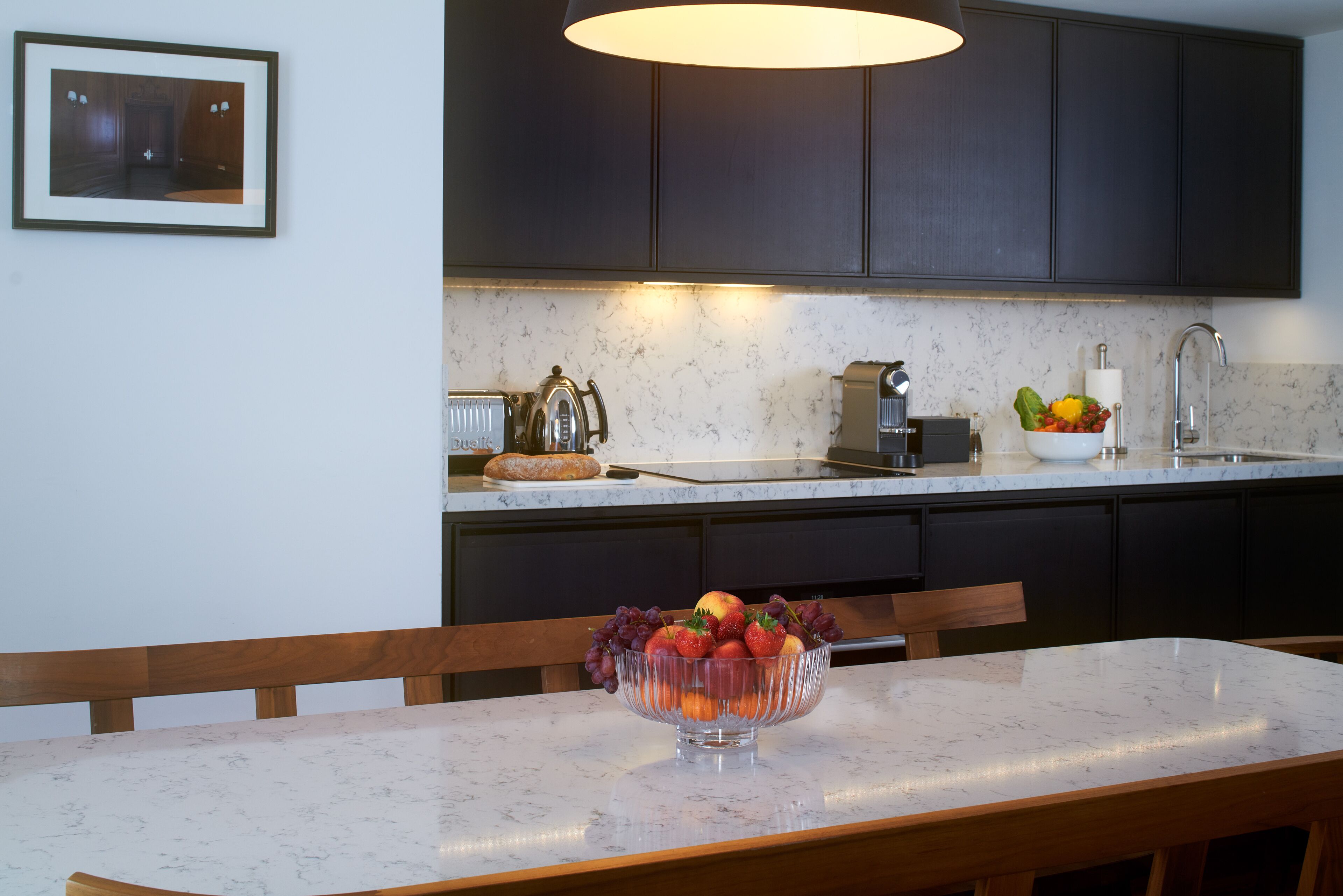 A modern kitchen with dark cabinets, a marble countertop, and a bowl of fruit on the table.