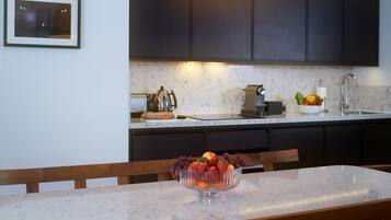 A modern kitchen with dark cabinets, a marble countertop, and a bowl of fruit on the table.