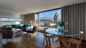 A modern living room with a large window, a sofa, a coffee table, and a dining area with chairs. - The Edinburgh Grand, a Luxury Collection Hotel, Edinburgh (Edinburgh)
