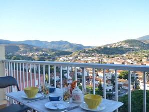 Uma varanda com uma mesa posta para dois, com vista para uma cidade com edifícios e colinas ao fundo.