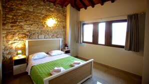 A bedroom with a stone wall, wooden ceiling beams, a bed with green bedding, a nightstand with a lamp, and a window with curtains. - Fruit Garden Villa (Kassandra)