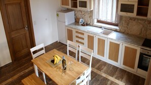 A compact kitchen with a wooden table, white chairs, a bench, a refrigerator, and a stone wall. - Fruit Garden Villa (Kassandra)
