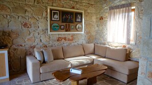 A stone-walled room with a beige sectional sofa, a wooden coffee table, and a wall-mounted picture frame with various items. - Fruit Garden Villa (Kassandra)
