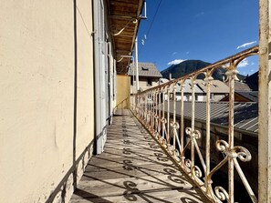 Varanda estreita com guarda-corrimão de metal, soalho de madeira e vista para edifícios e montanhas ao fundo.
