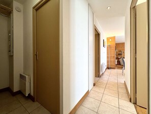 A hallway with a closed door, a radiator, and a tiled floor leading to a room with a wooden wall and a chair.