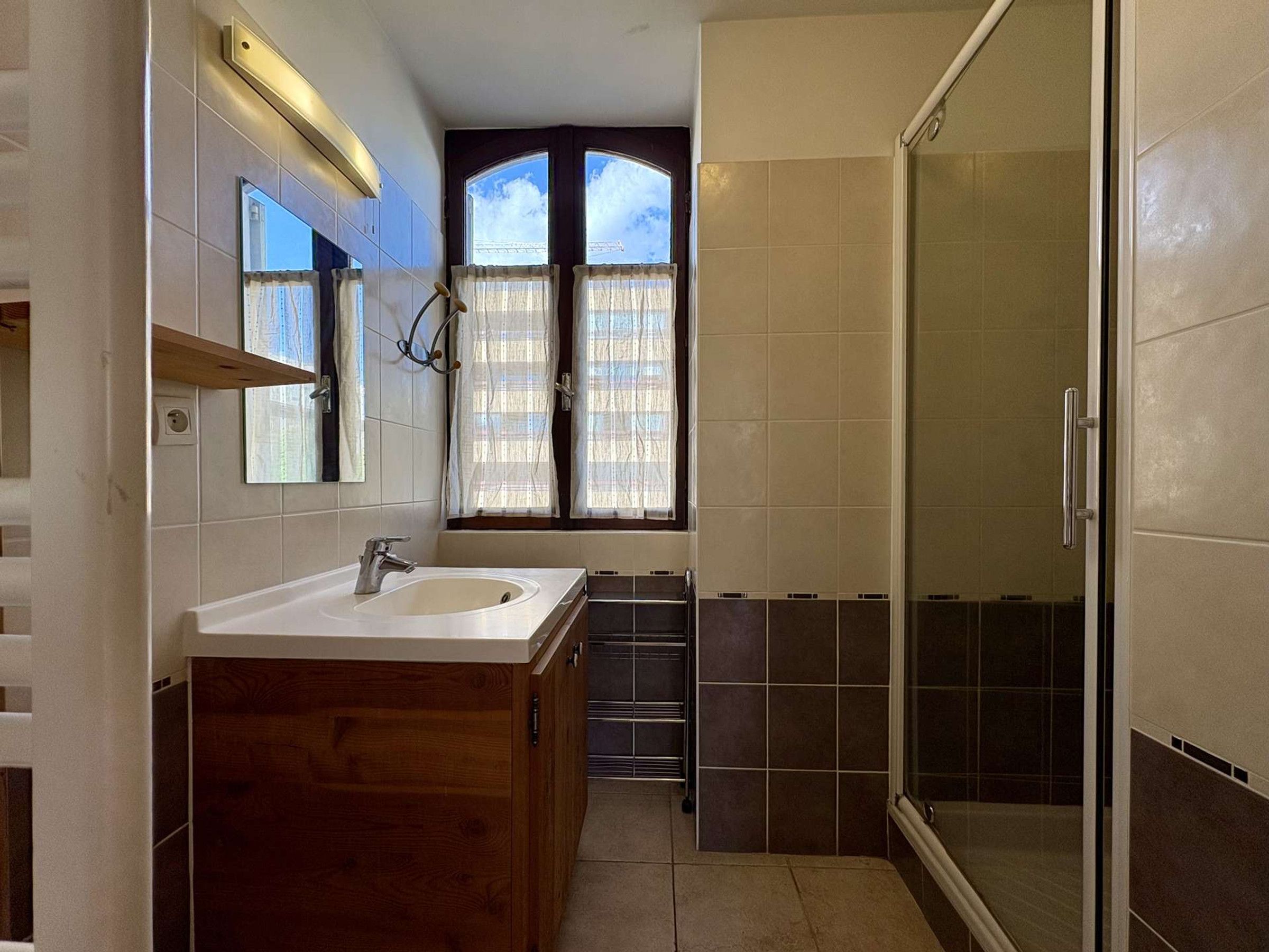 A bathroom with a wooden vanity, a sink, a mirror, a window with curtains, and a shower area.