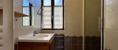 A bathroom with a wooden vanity, a sink, a mirror, a window with curtains, and a shower area.