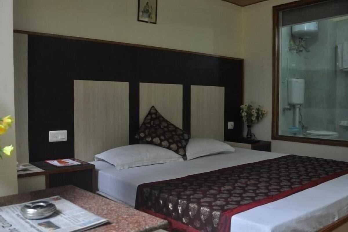A hotel room with a bed, a bedside table, a nightstand, a window, and a vase with flowers.