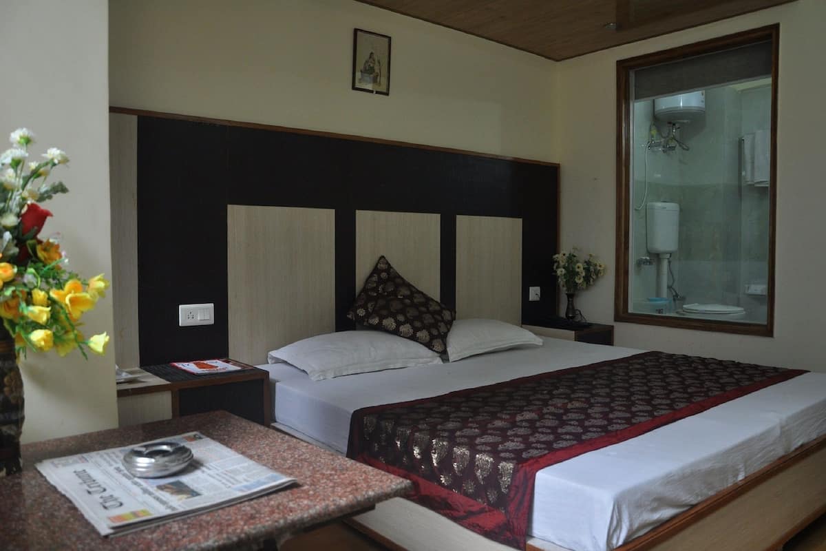 A hotel room with a bed, a bedside table with a newspaper, a vase with flowers, and a window with a view of a bathroom.
