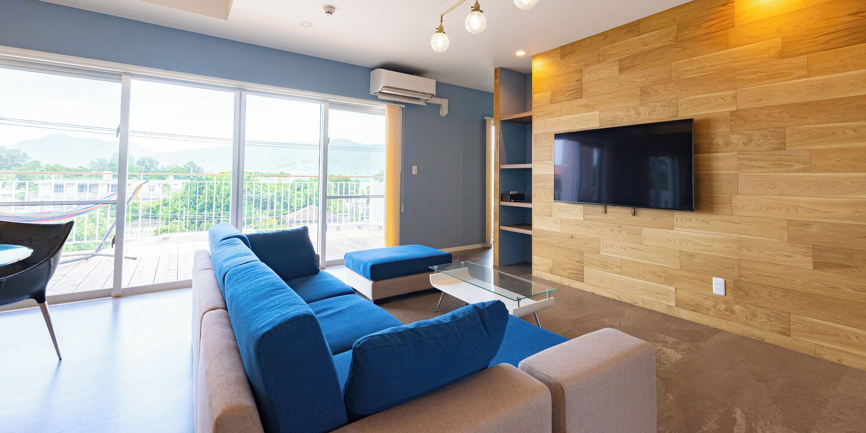 A modern living room with a large TV mounted on a wooden wall, a sofa with blue cushions, and a glass coffee table.