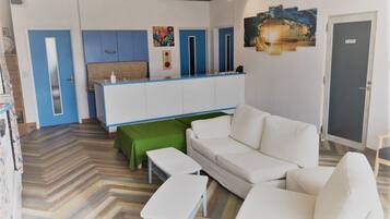 A modern hotel room with a white sofa, a green ottoman, a white table, and a kitchenette with blue cabinets.