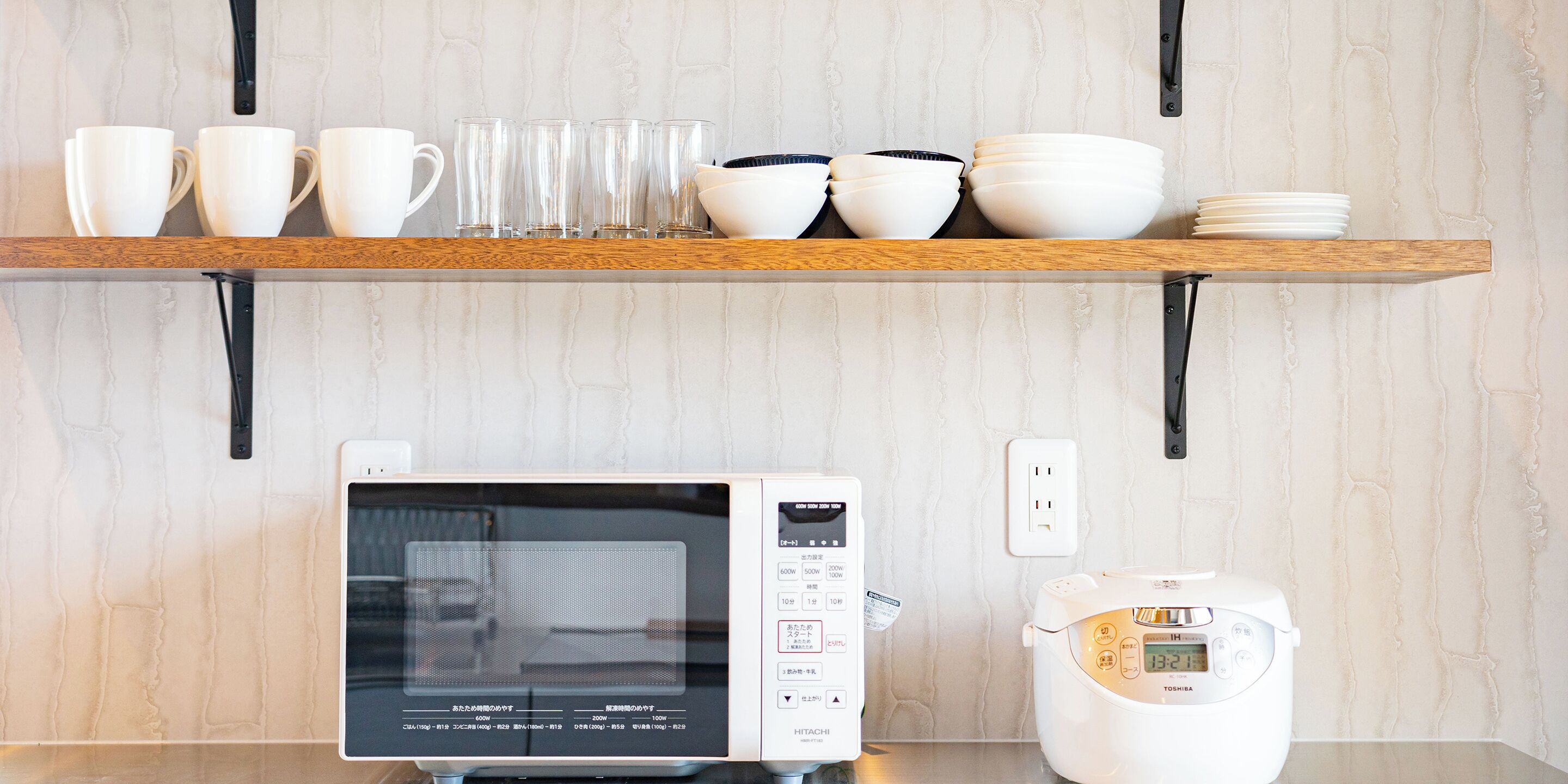 Una cocina con microondas y cocedor de arroz sobre la encimera, un estante arriba con tazas y cuencos, y una pared con textura.