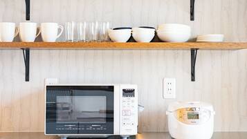 Una cocina con microondas y cocedor de arroz sobre la encimera, un estante arriba con tazas y cuencos, y una pared con textura.
