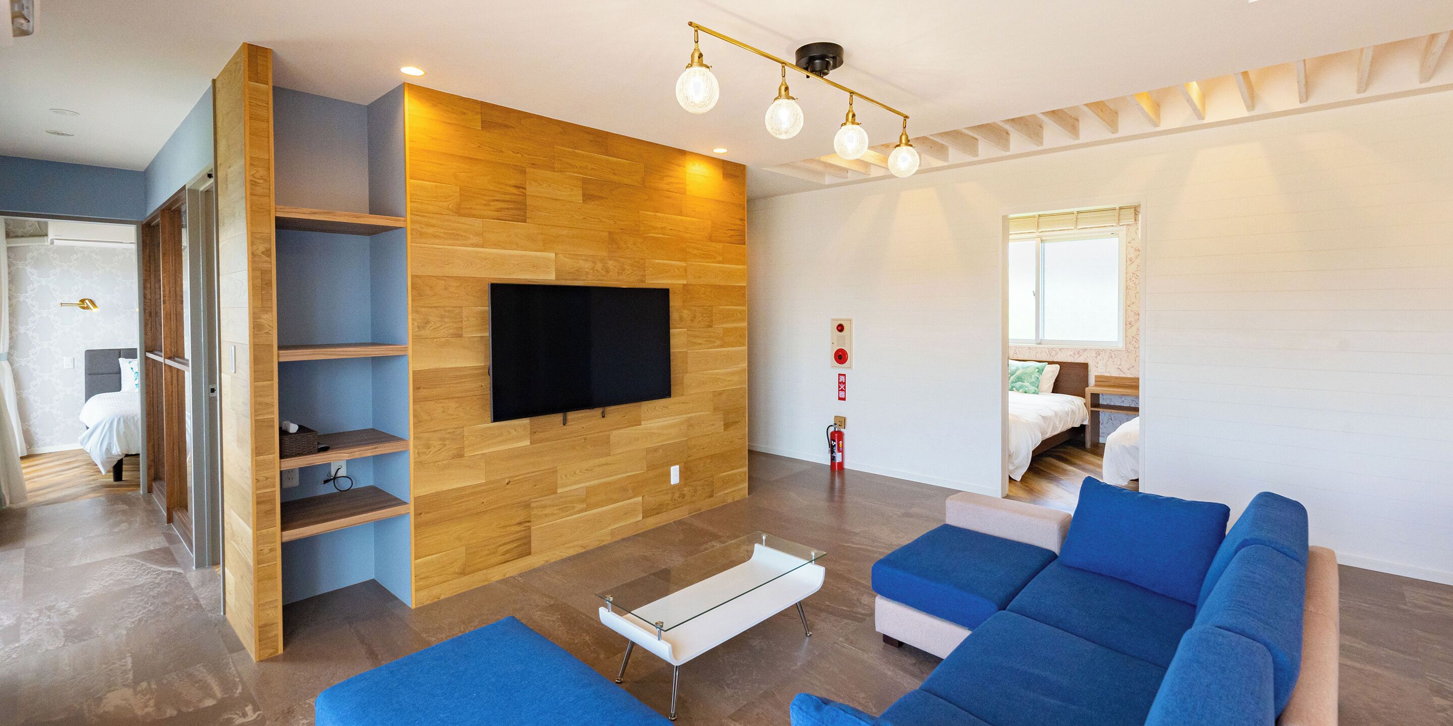 A modern living room with a flat-screen TV mounted on a wooden wall, a blue sofa, a glass coffee table, and a wooden shelving unit.