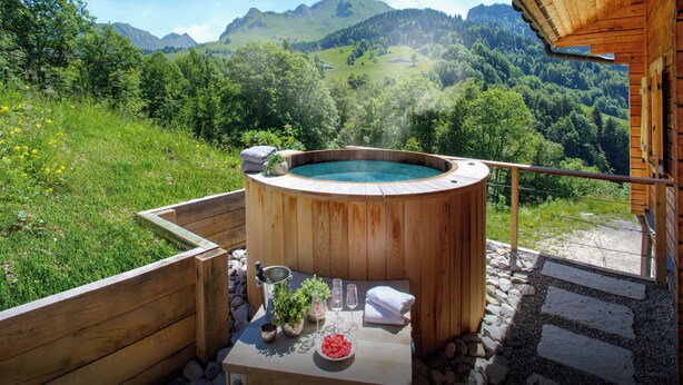 Un jacuzzi de madera con una mesa para dos, rodeado por una terraza de madera y un camino de piedra, con vistas a un valle exuberante y montañas.
