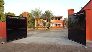 A gated entrance to a building with a red and white facade, surrounded by palm trees and a dirt ground.
