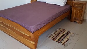 A wooden bed with a purple bedspread and white pillows, flanked by wooden nightstands with drawers, on a tiled floor with a striped rug.