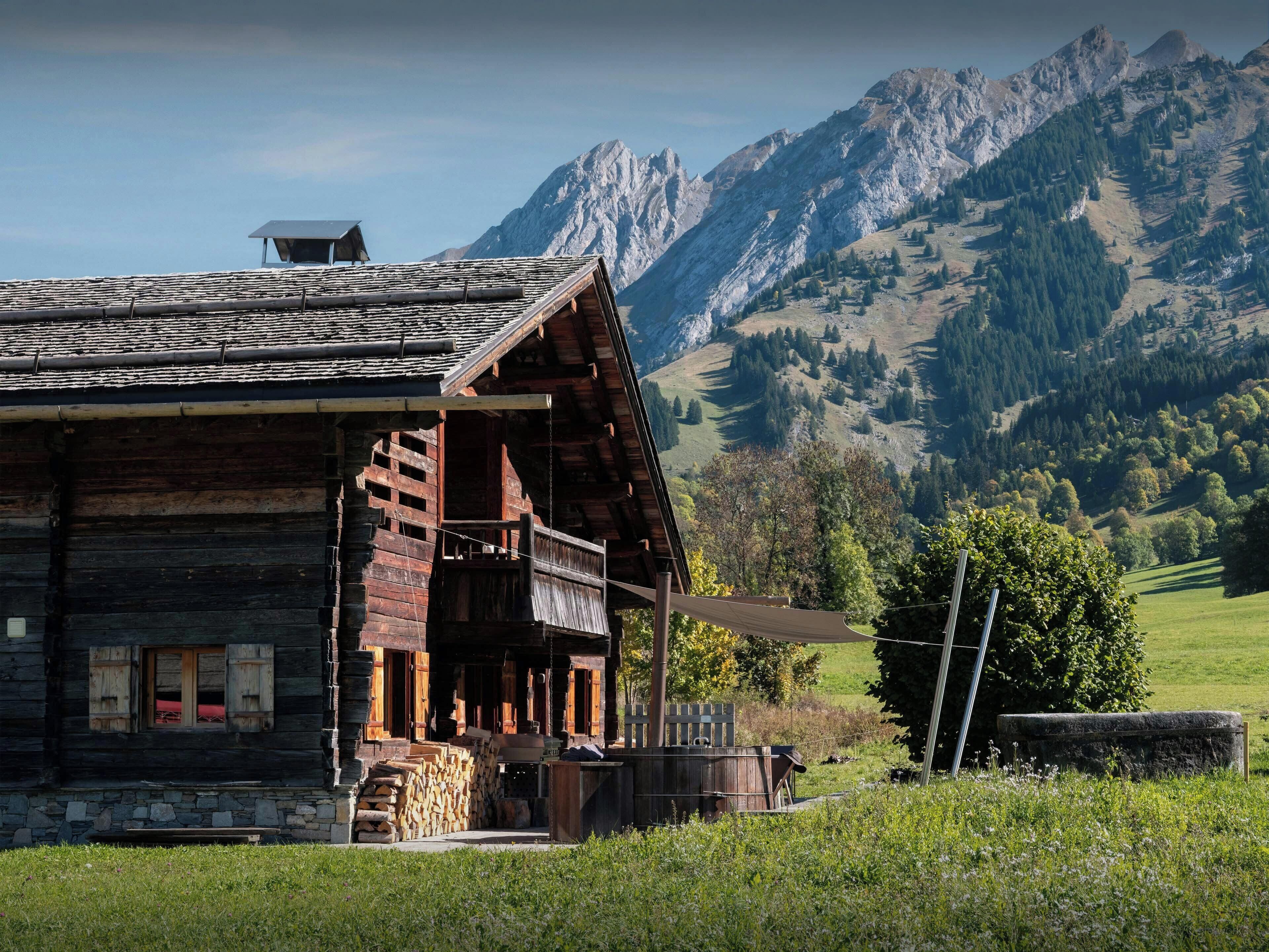 A rustic wooden cabin with a balcony, surrounded by a lush green landscape and mountains in the background.
