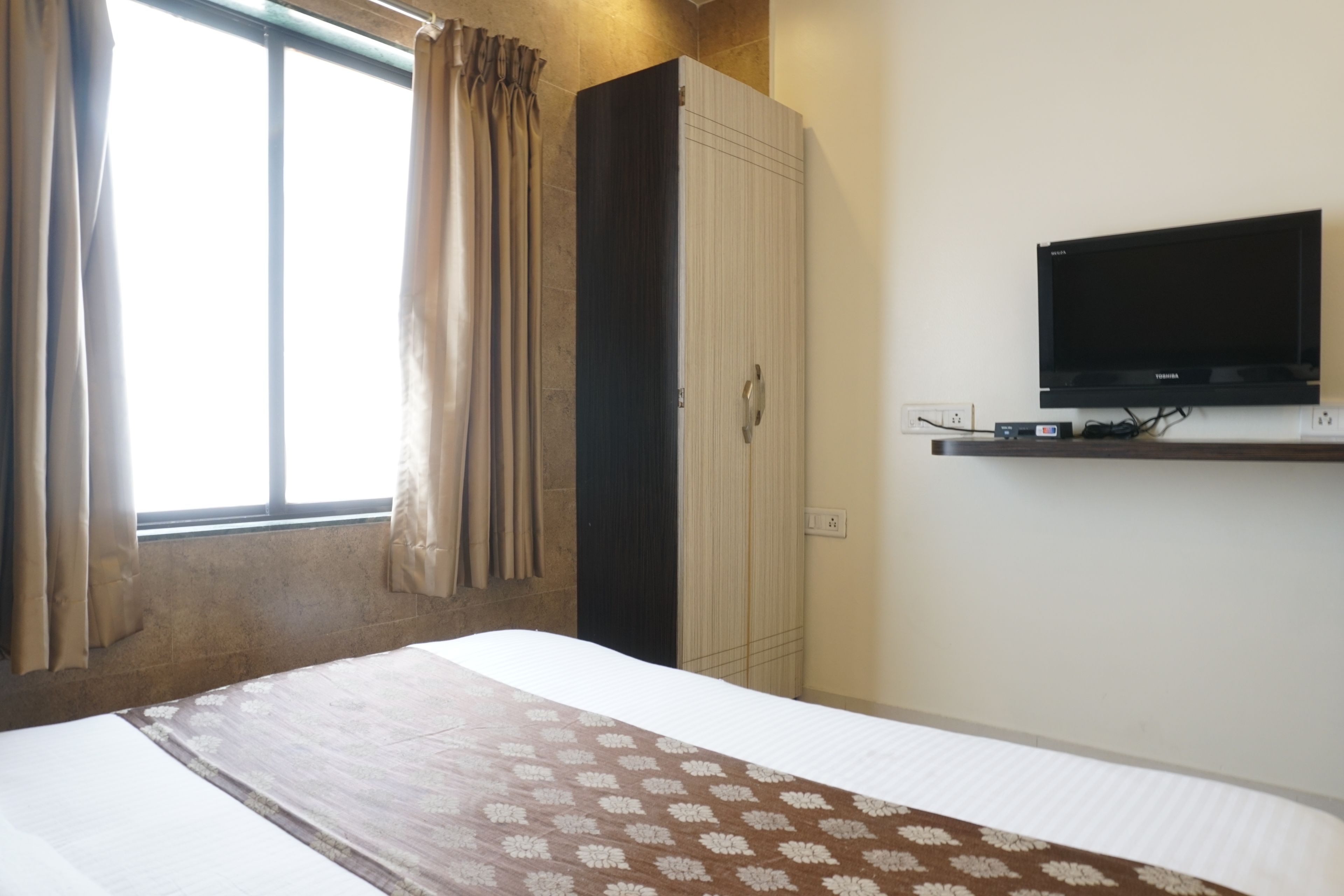 Habitación de hotel con cama, armario, televisor montado en la pared y ventana con cortinas.