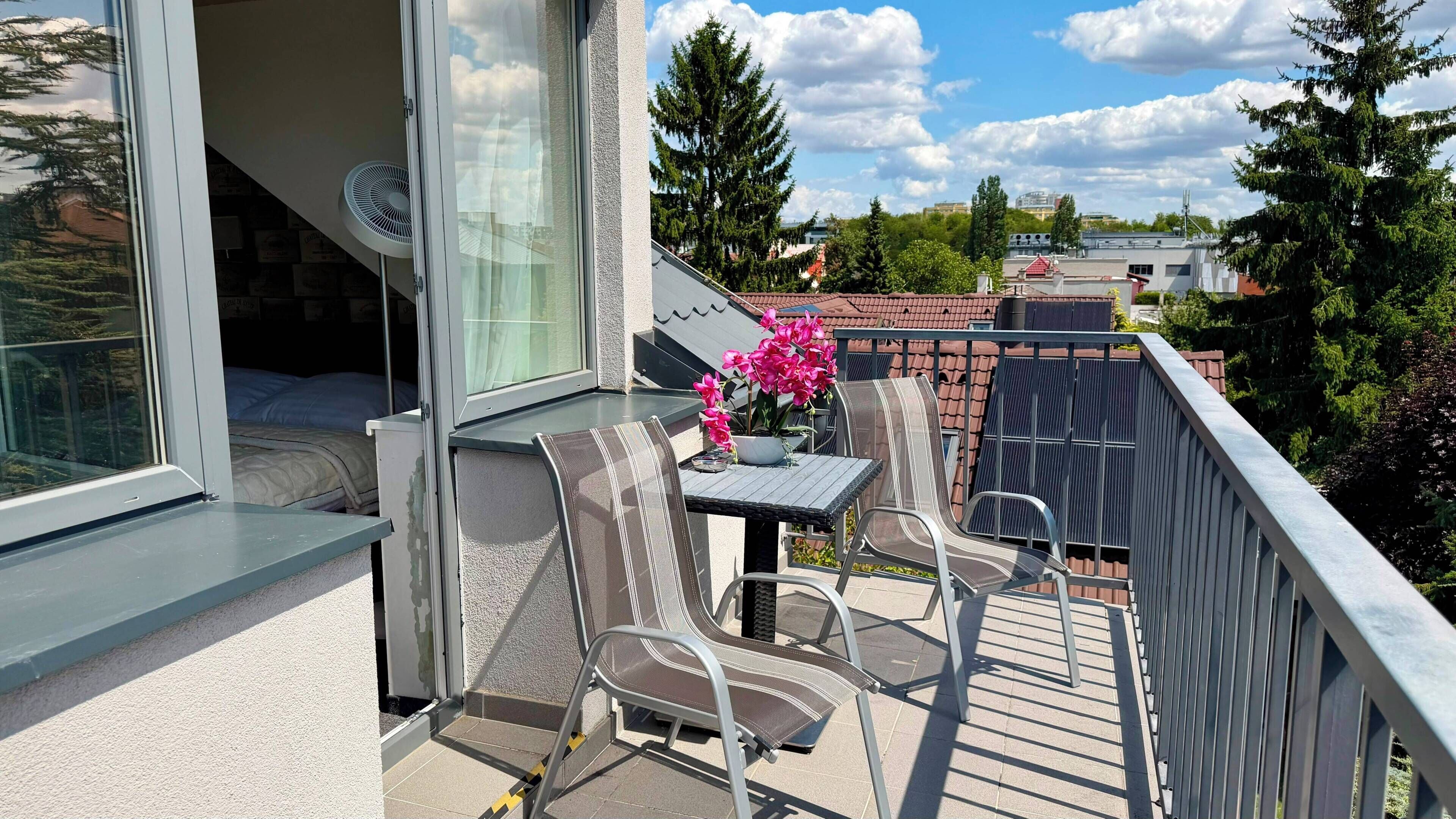 Un balcón con una mesa y sillas, una planta en maceta y vistas de los edificios y árboles circundantes.