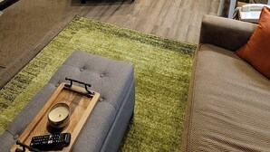 Un salon moderne avec un canapé, une table basse sur laquelle reposent des télécommandes, et un fauteuil à motifs. On aperçoit, au fond, une cuisine équipée d’appareils électroménagers en acier inoxydable.