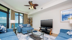 Una sala de estar moderna con un sofá azul, una mesa de centro de vidrio, un televisor de pantalla plana y un balcón con vista al océano.