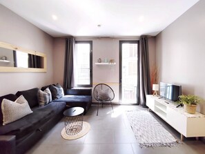 Un salon moderne avec un canapé, une table basse ronde, un téléviseur, une plante et une vue sur l’extérieur.