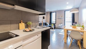 A modern kitchen with a built-in dishwasher, a dining area with a wooden table and chairs, and a living space with a sofa and a window. - Bo&Co Apartments Sitges (Sitges)