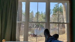 Une pièce avec une grande fenêtre donnant sur un lac, un lit recouvert d’une couette blanche, et une paire de pieds reposant sur le rebord de la fenêtre.