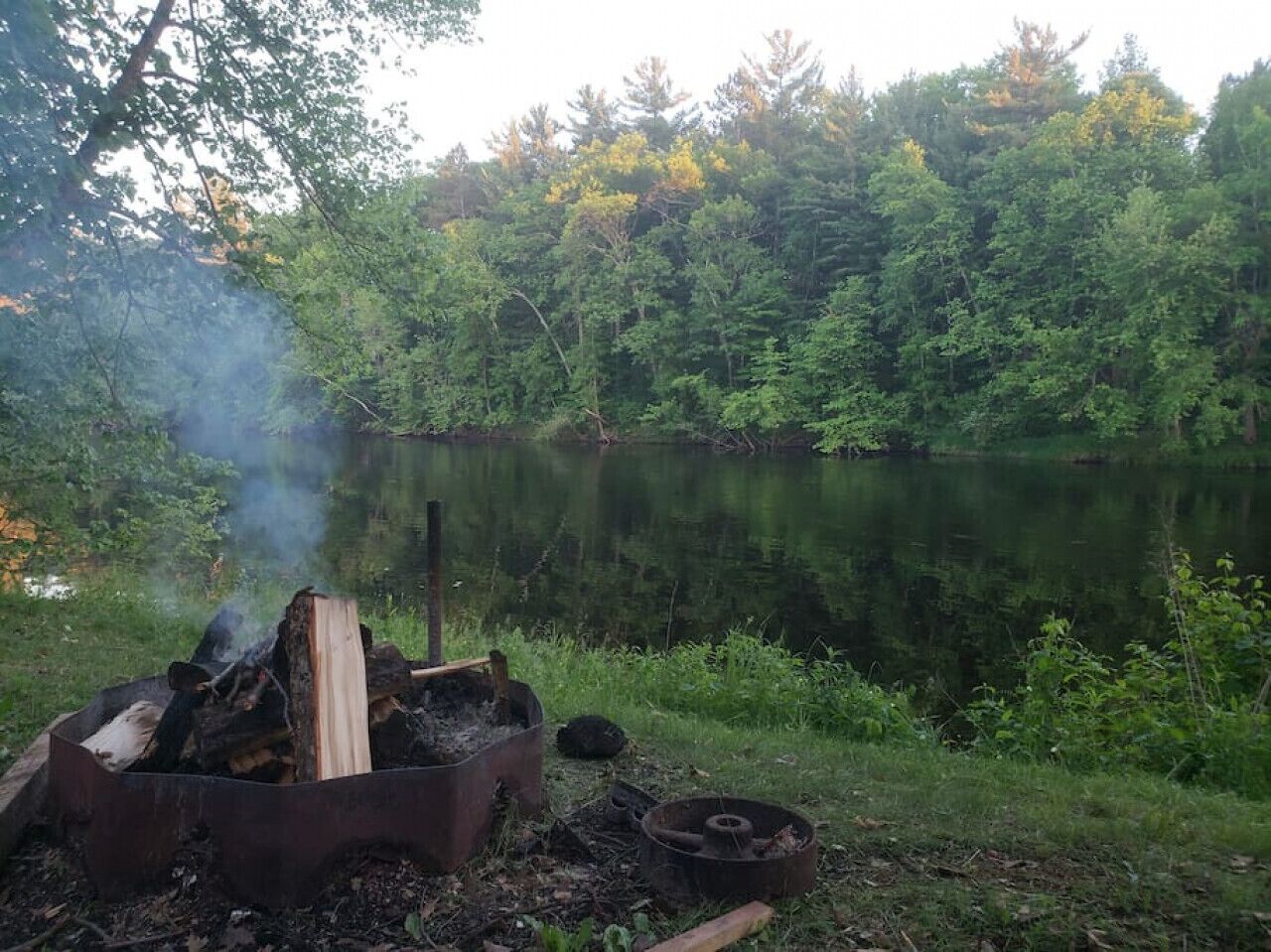 A campfire with logs and a metal container, surrounded by a forest and a calm lake.