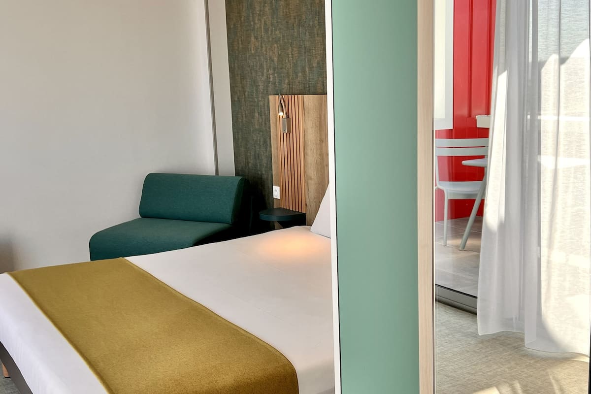 A hotel room with a bed, a green chair, a small table, and a red and white chair near a window with sheer curtains.