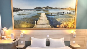A hotel room with a large bed, two bedside tables, a desk, and a painting of a coastal landscape.