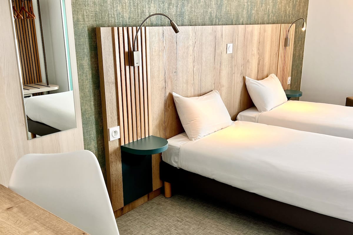 A hotel room with two beds, a wooden headboard, bedside tables, a mirror, and a small desk with a chair.