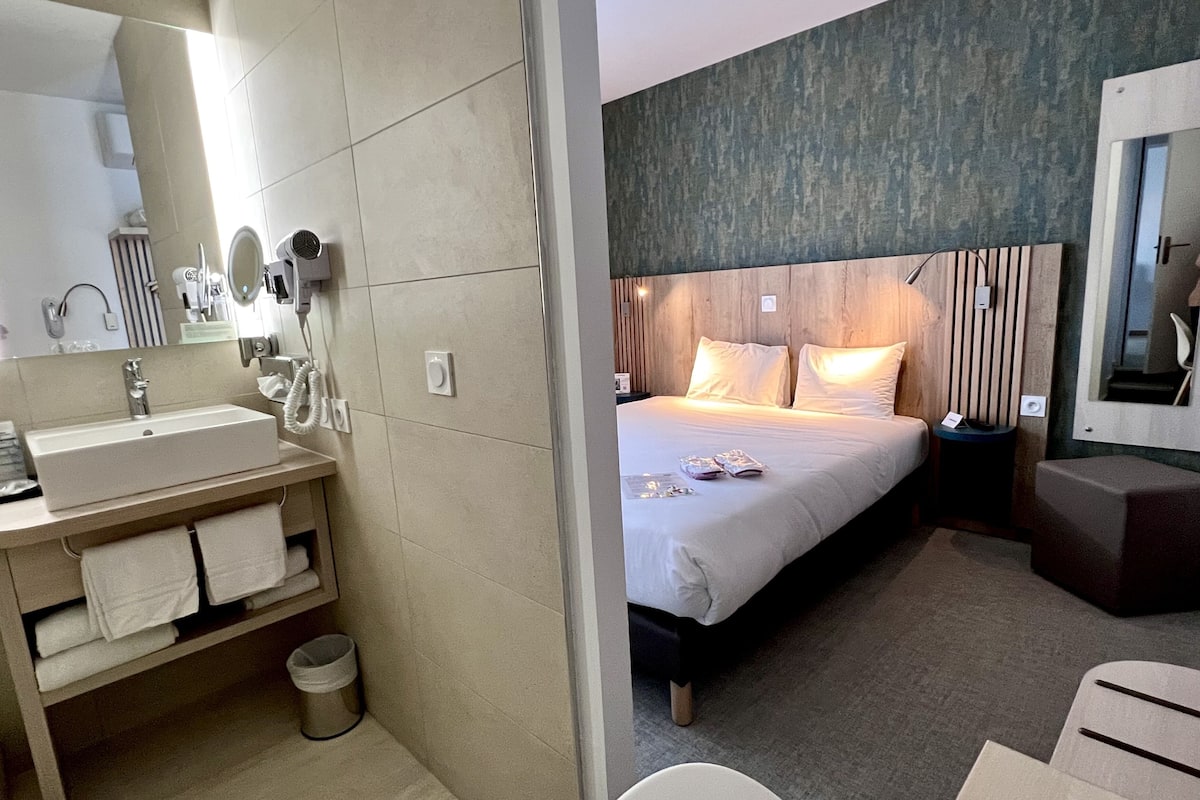 A modern hotel room with a large bed, a bedside table, a nightstand, a bathroom with a sink, mirror, and towel rack.
