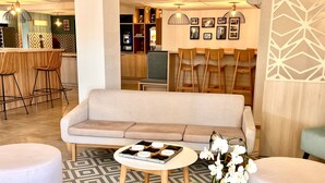 A modern lobby with a sofa, two round tables, and chairs. There are decorative plants and a coffee machine in the background.