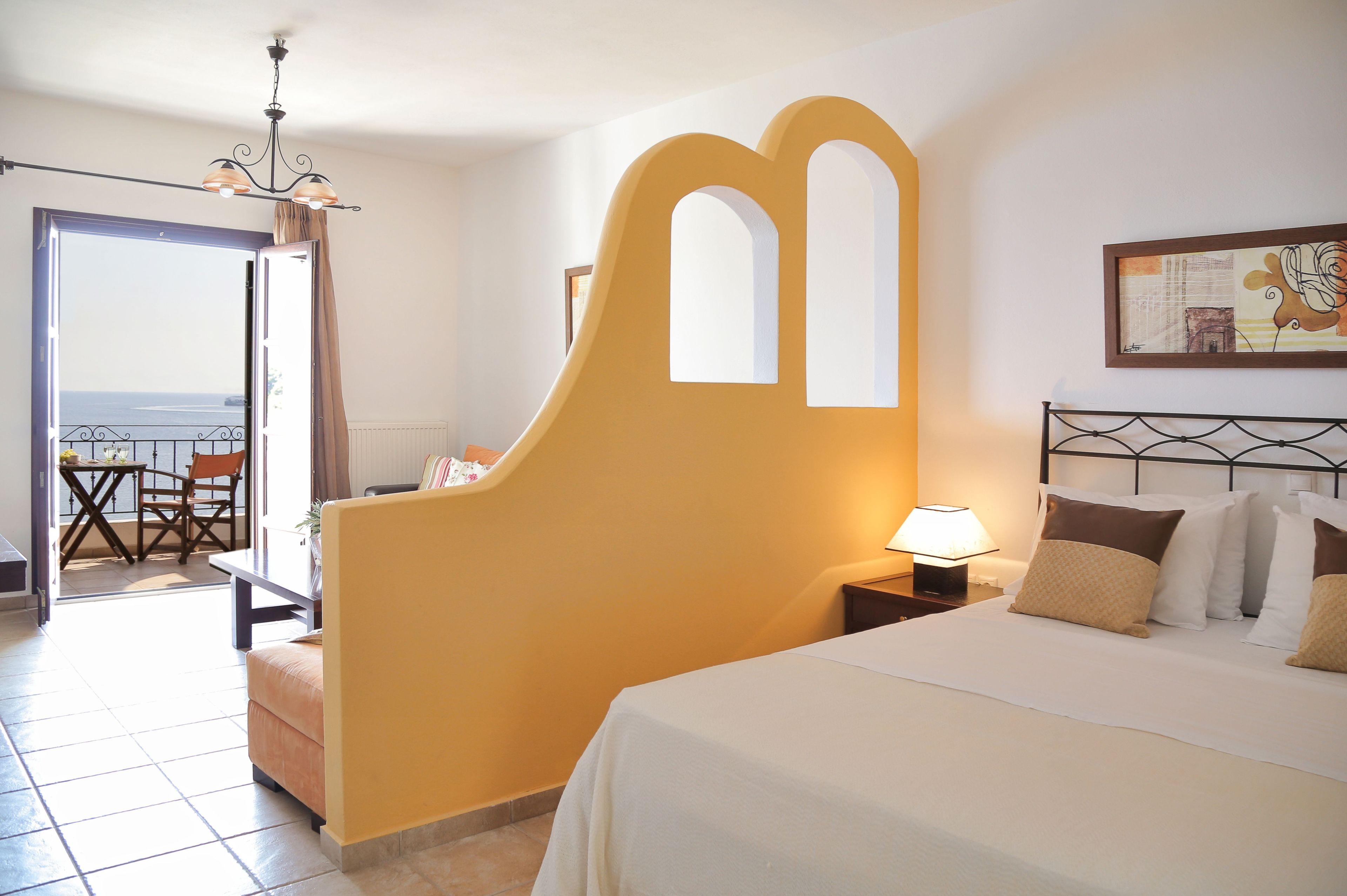 Een hotelkamer met een bed, nachtkastje, lamp, stoel, tafel en uitzicht op de zee door een open deur.