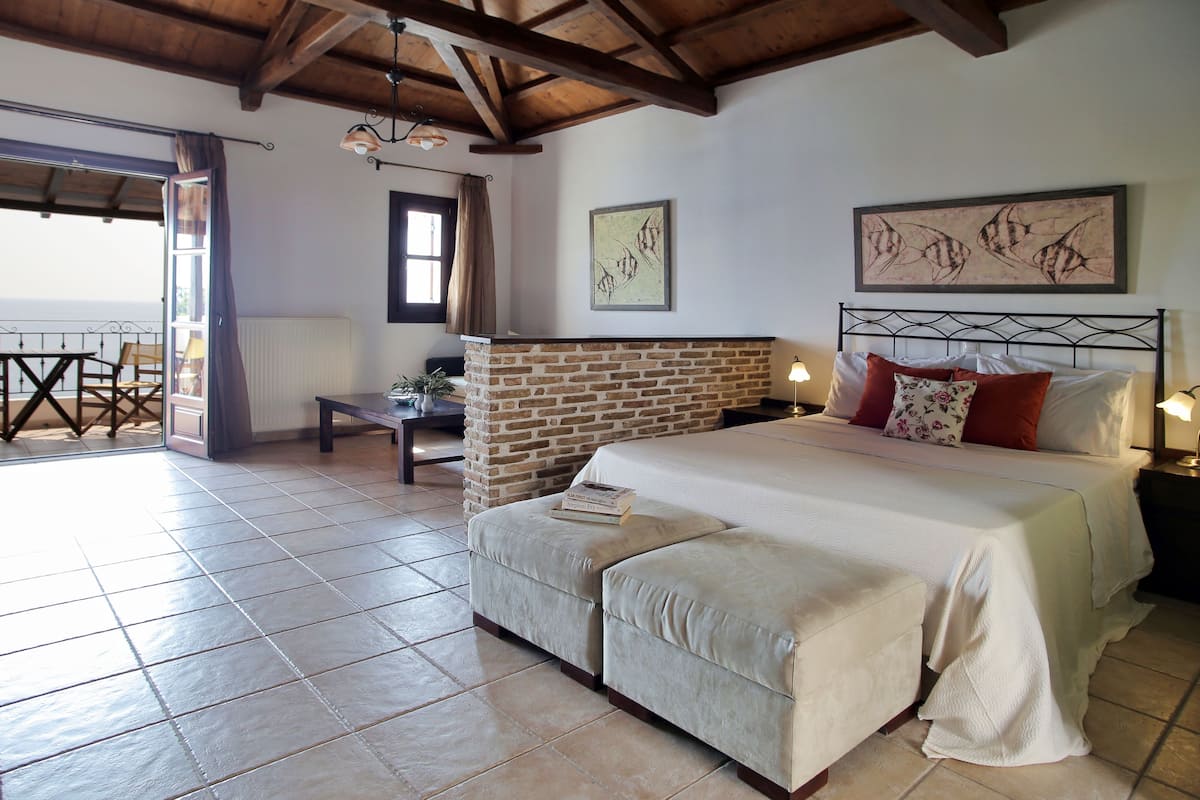 A bedroom with a stone fireplace, a bed with white bedding, and a view of the outside.