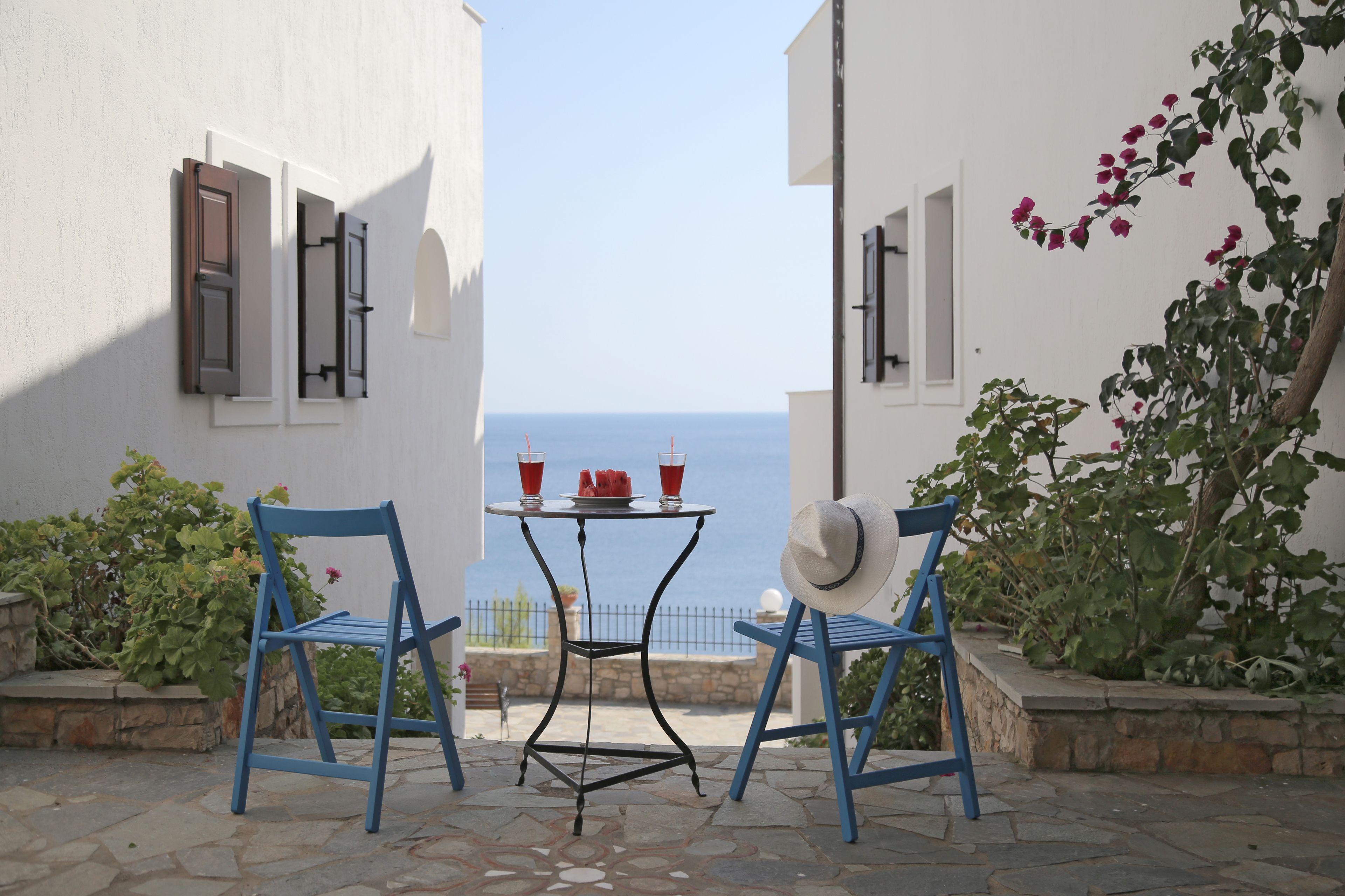 Una terraza con dos sillas azules, una mesa con dos tazas rojas y un plato de fruta, y vistas al mar.