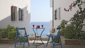 Una terraza con dos sillas azules, una mesa con dos tazas rojas y un plato de fruta, y vistas al mar.