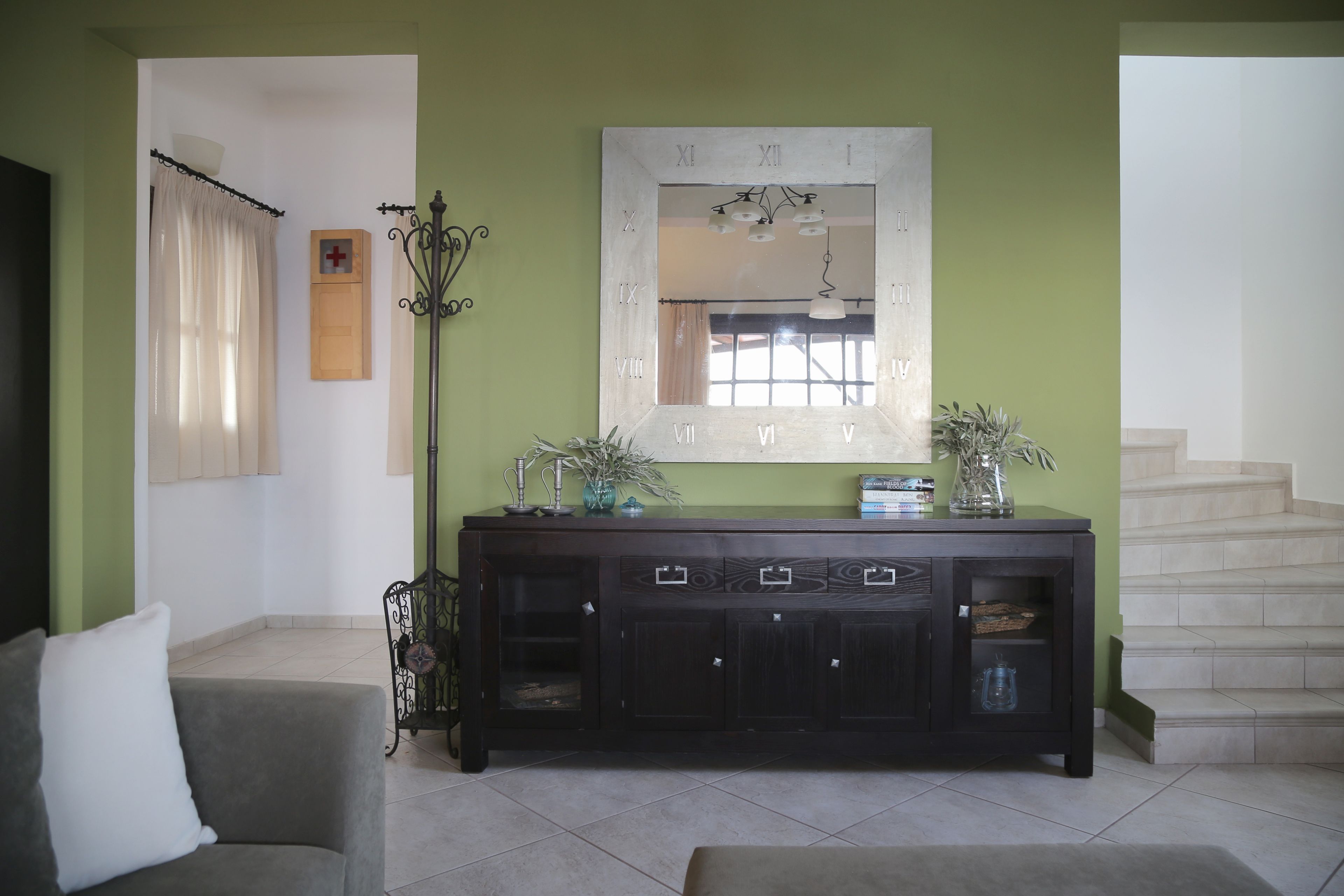 Una sala de estar con una consola de madera oscura, un espejo grande con números romanos, una escalera y una pared verde.
