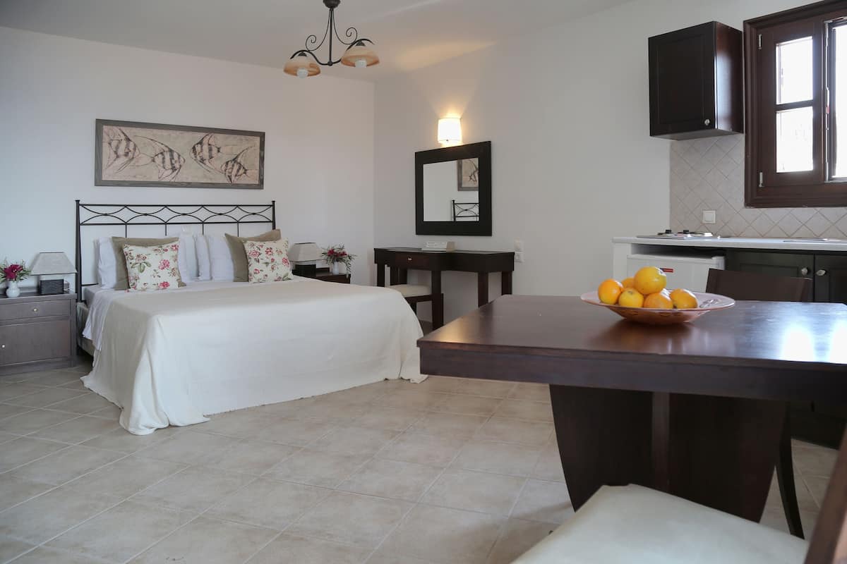 A bedroom with a bed, bedside table, and a nightstand. There is a dining table with a bowl of fruit in the kitchen area.