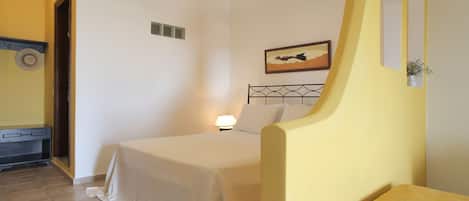 Un dormitorio con cama, lámpara de noche, una mesita y un cuadro colgado en la pared.