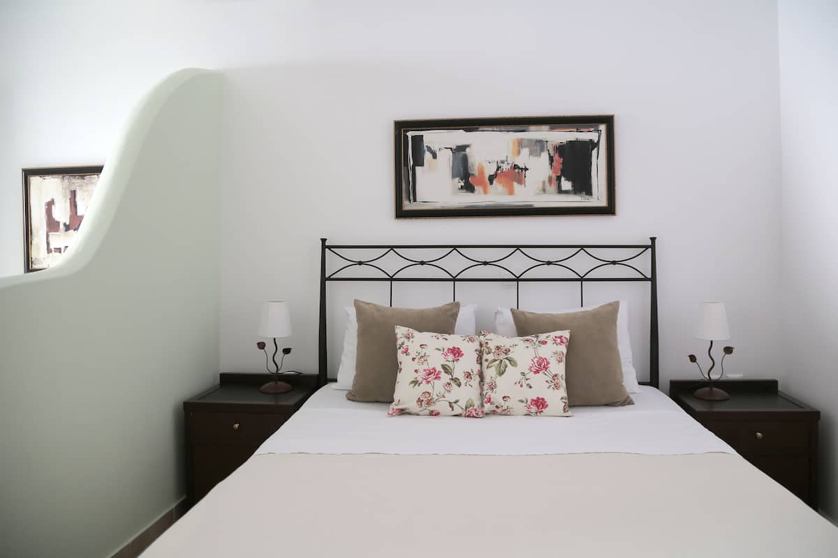 A neatly arranged bedroom with a bed, two nightstands, a wall-mounted abstract painting, and a mirror reflecting part of the room.