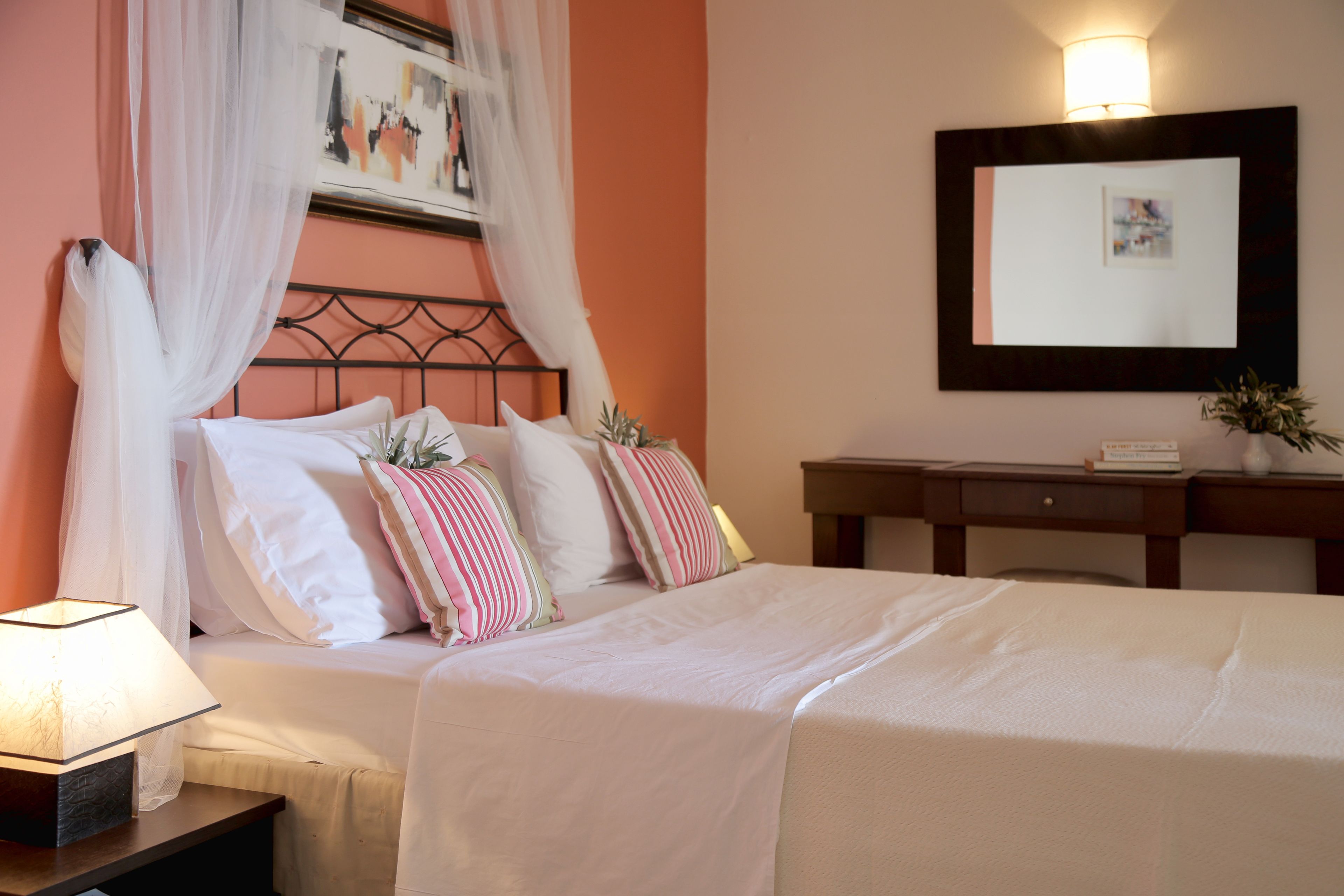 A neatly made bed with white linens, a bedside lamp, and a nightstand. There is a wooden dresser with a mirror above it and a framed picture on the wall.