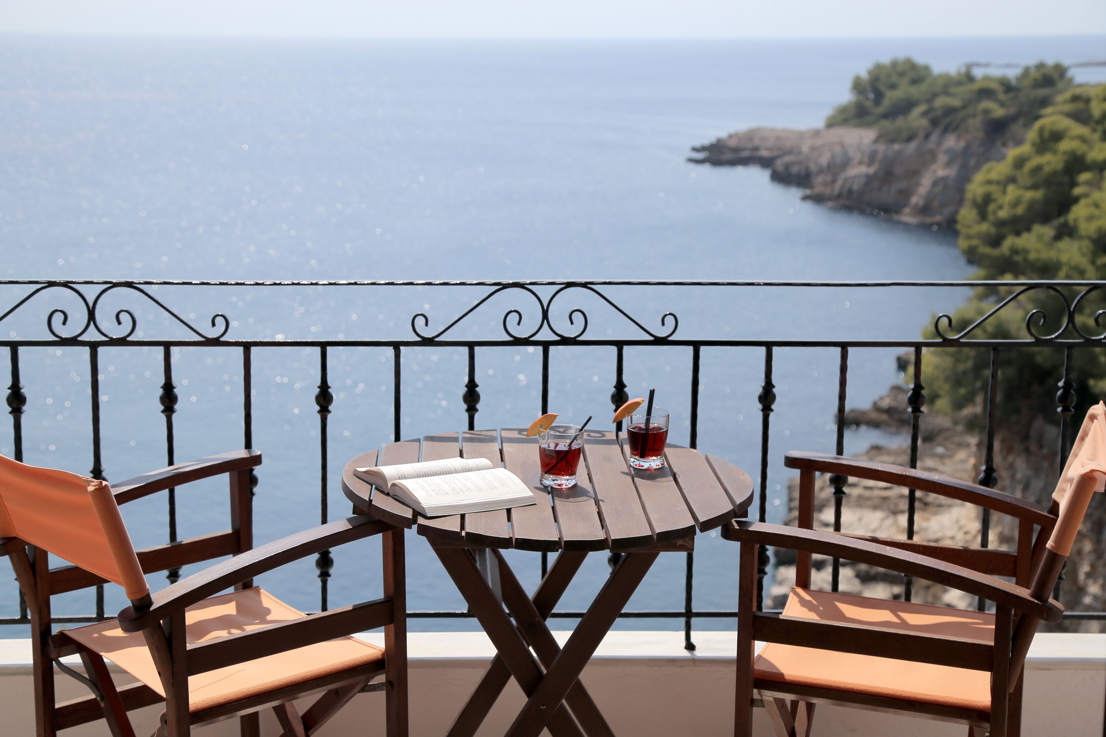 A wooden table with two chairs, a book, and two drinks on a balcony overlooking a coastal view.