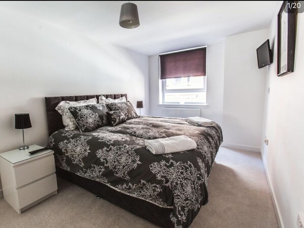 A bedroom with a large bed, a bedside table, a lamp, a window with a blind, and a wall-mounted TV.