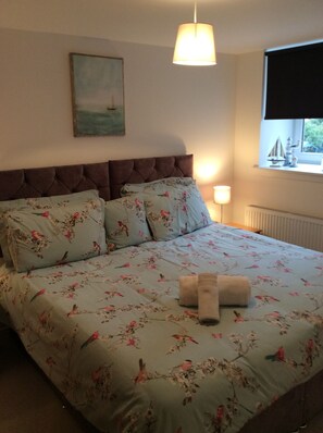 A bedroom with a bed, a headboard, a painting on the wall, a window with a view, and a lamp.