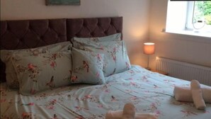 A bedroom with a tufted headboard, floral bedding, a painting on the wall, a bedside lamp, and a window with a view. - Mossie Apartment (Ayr)