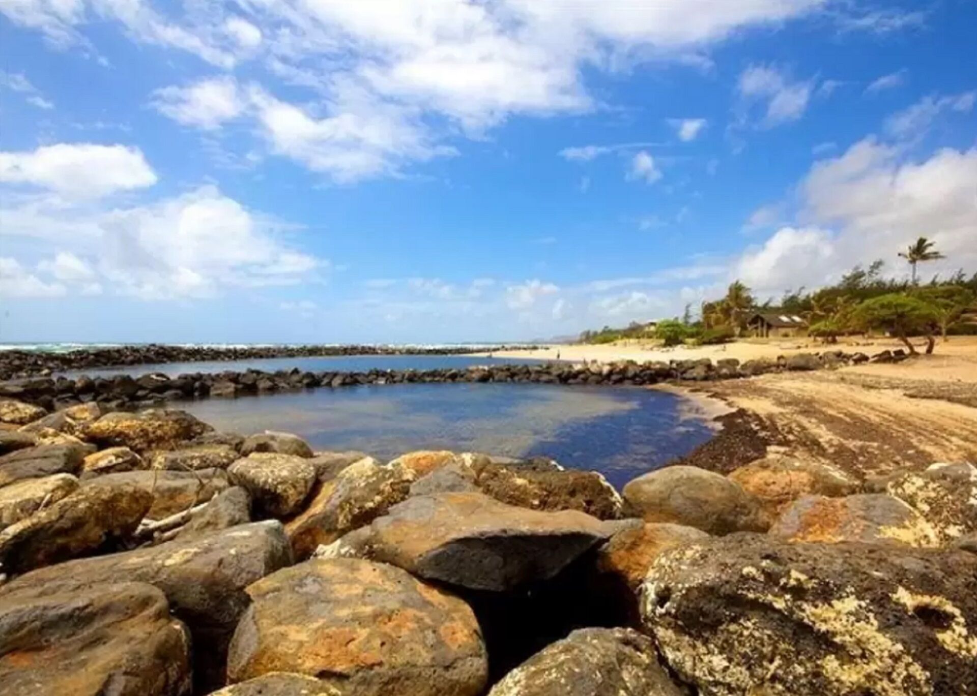A rocky shoreline with a calm sea, a clear sky, and a distant view of a beach with vegetation and a structure.