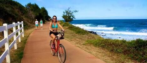 A paved pathway alongside a coastal area with a white fence on one side and the ocean on the other.